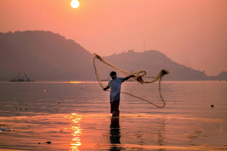 a man that is standing in the water with a net, inspired by Steve McCurry, pexels contest winner, sun setting, hindu art, fishing, warm coloured