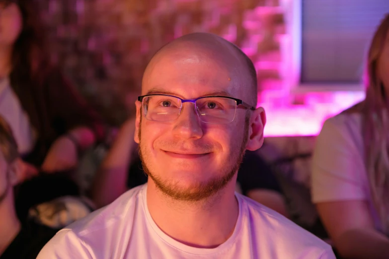 a man with glasses sitting in front of a group of people, by Adam Marczyński, reddit, headshot profile picture, glowing, slight friendly smile, pale head