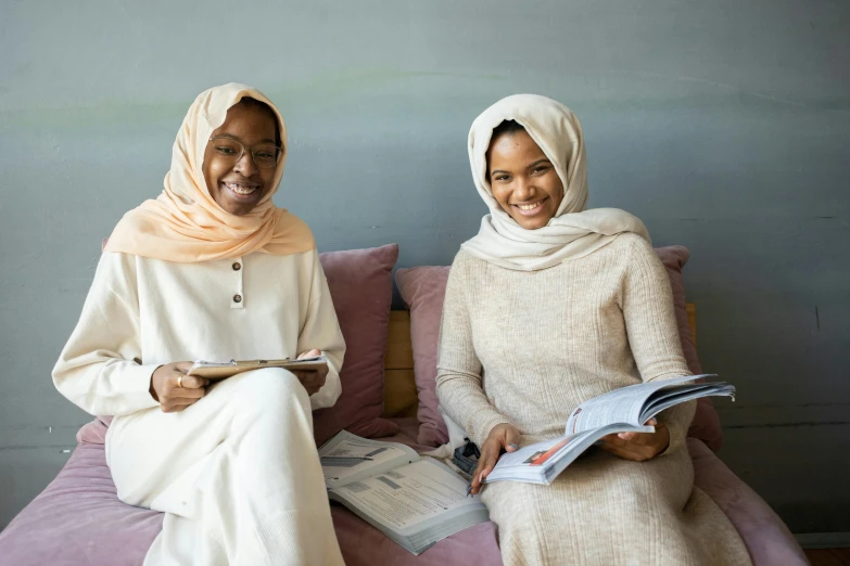 a couple of women sitting on top of a pink couch, a portrait, trending on unsplash, hurufiyya, white hijab, holding books, with brown skin, swedish