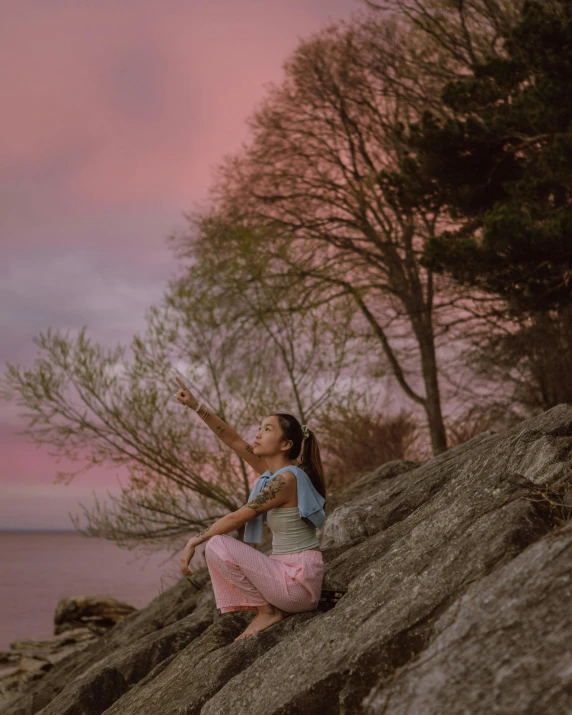 a woman sitting on top of a rock next to a body of water, pink skies, next to a tree, thinking pose, waving