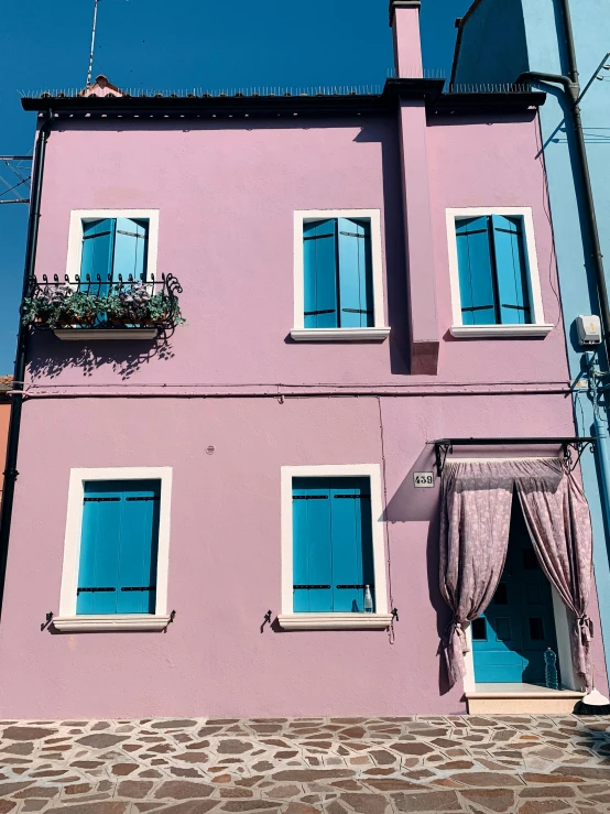 a pink building with blue windows on a cobblestone street, inspired by Ricardo Bofill, pexels contest winner, mauve and cyan, sunny bay window, italy, wires hanging across windows