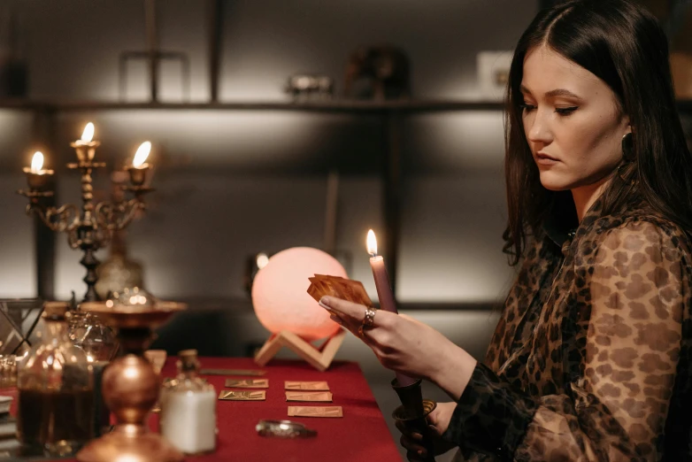 a woman sitting at a table holding a candle, a hologram, trending on pexels, wizard shuffling cards, wearing red sorcerers robes, concentration, apothecary