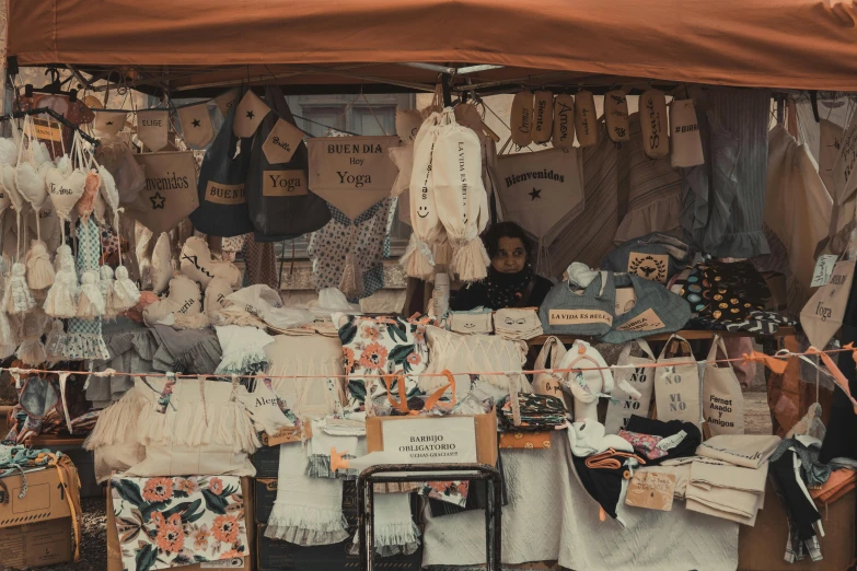 a woman standing in front of a tent with clothes on it, by Emma Andijewska, pexels contest winner, folk art, lots of signs and shops, market stalls, sitting down, brown