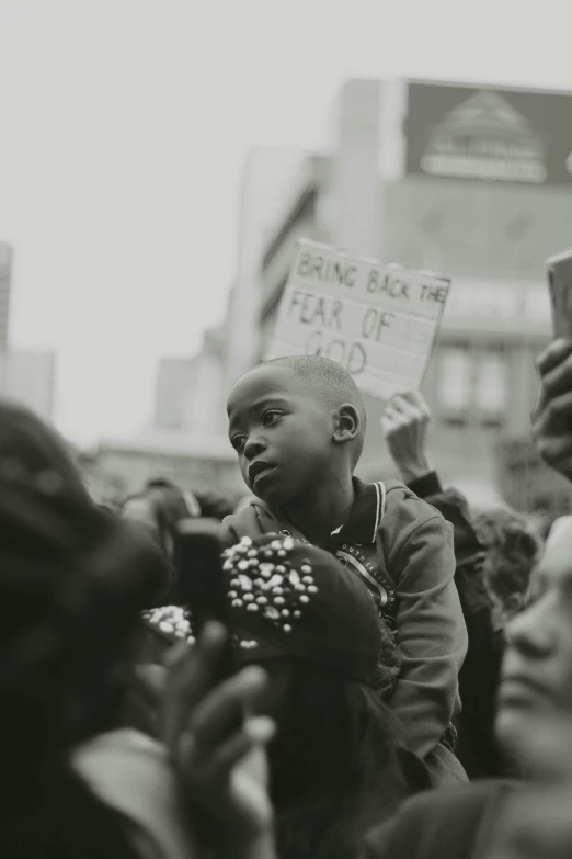 a black and white photo of a man holding a sign, trending on unsplash, black arts movement, with a kid, crowds of people praying, boy with neutral face, young thug