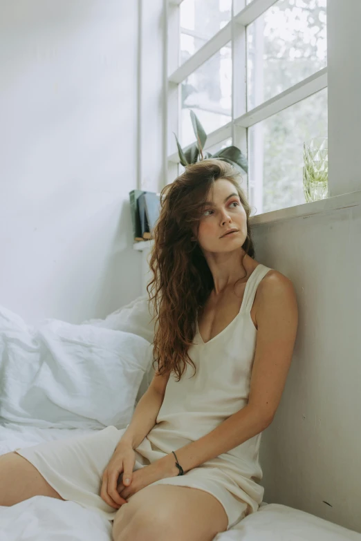 a woman sitting on top of a bed next to a window, inspired by Elsa Bleda, renaissance, no makeup wavy hair, looking to the side off camera, in front of white back drop, contemplative