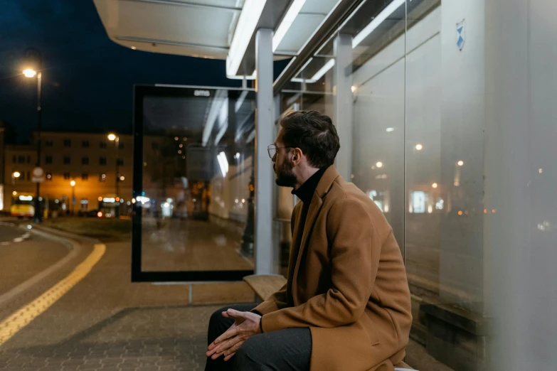 a man sitting on a bench at a bus stop, by Adam Marczyński, pexels contest winner, light brown coat, avatar image, very reflective, transparent