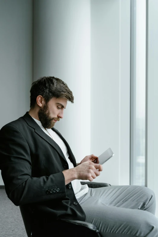 a man sitting in a chair looking at his cell phone, well trimmed beard, big windows, wearing a black blazer, holding notebook