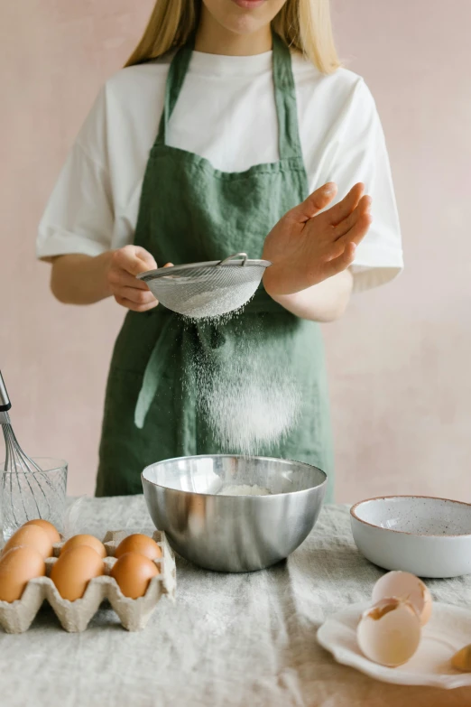 a woman standing in front of a table with eggs and a whisk, trending on pexels, flour dust, bowl, wearing an apron, greens)