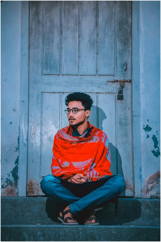 a man sitting on the ground in front of a door, inspired by Nabil Kanso, pexels contest winner, he‘s wearing a red scarf, jewish young man with glasses, assamese aesthetic, full body hero