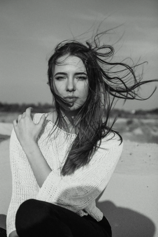 a black and white photo of a woman sitting on the beach, hair blowing in the wind, perfect face ), wearing a white sweater, trending on imagestation