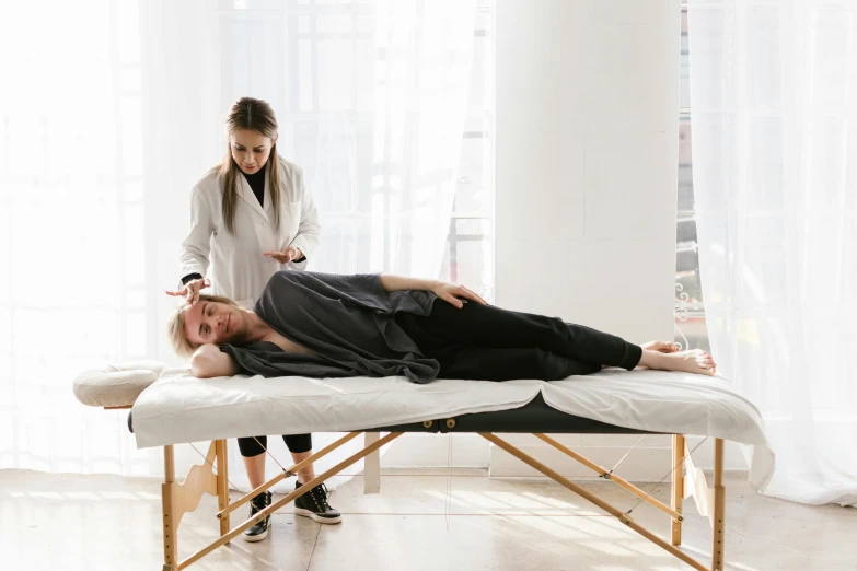 a man laying on a massage table next to a woman, wearing a grey robe, ouchh and and innate studio, on a white table, supportive