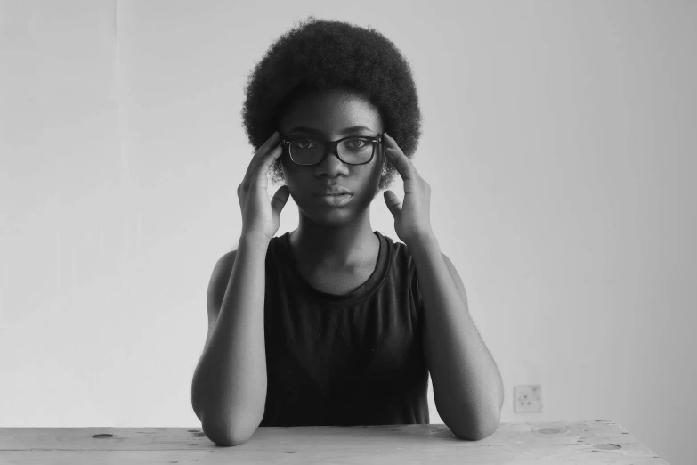 a woman sitting at a table with her hands on her head, a black and white photo, by Lily Delissa Joseph, pexels, afrofuturism, with square glasses, symmetry!! portrait of a woman, black teenage boy, with afro