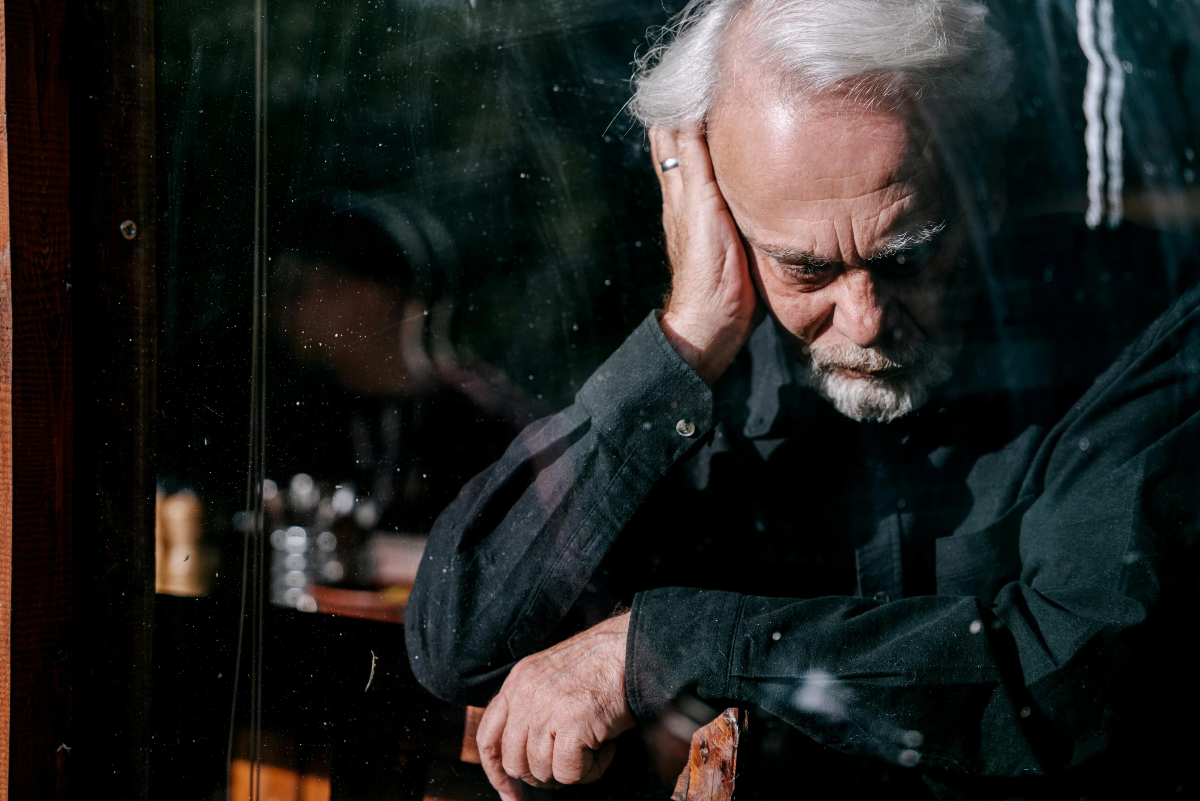 a man sitting in front of a window with his hand on his head, pexels contest winner, precisionism, white hair and beard, neil young, a man wearing a black jacket, puzzling