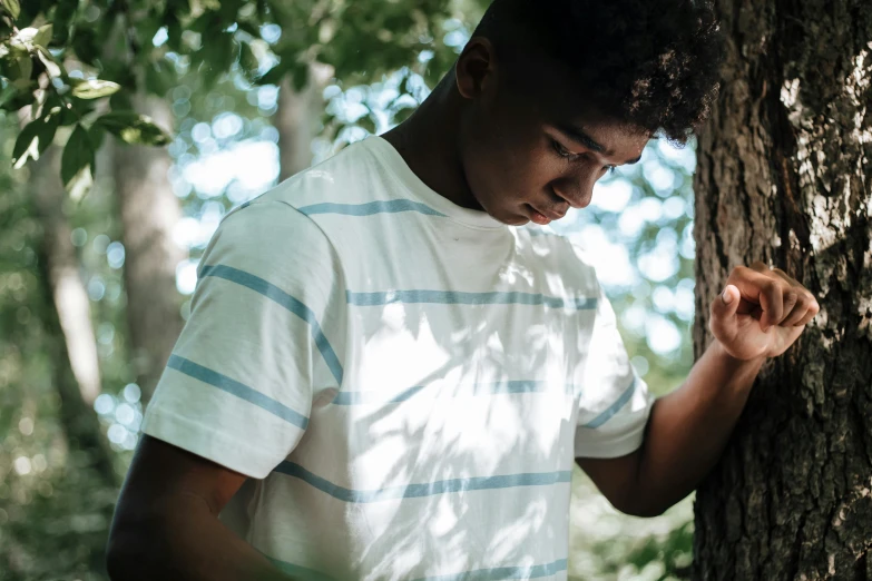 a young man standing next to a tree, by Everett Warner, trending on pexels, happening, black teenage boy, shy looking down, dressed in a white t shirt, wearing stripe shirt