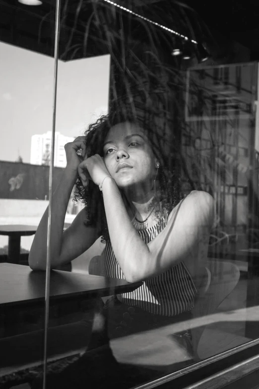 a black and white photo of a woman sitting at a table, a black and white photo, pexels, aida muluneh, leaning against the window, concerned, in the sun