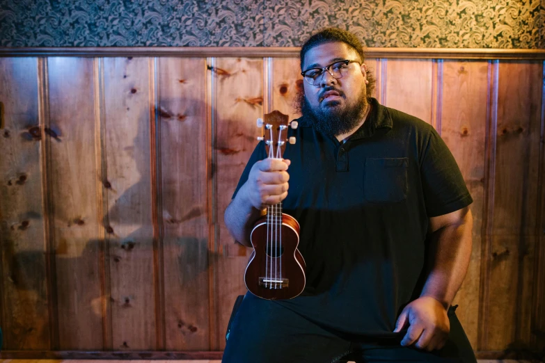 a man holding a guitar in front of a wooden wall, by Matt Cavotta, hurufiyya, similar to hagrid, jontron, standing in a dimly lit room, ukulele