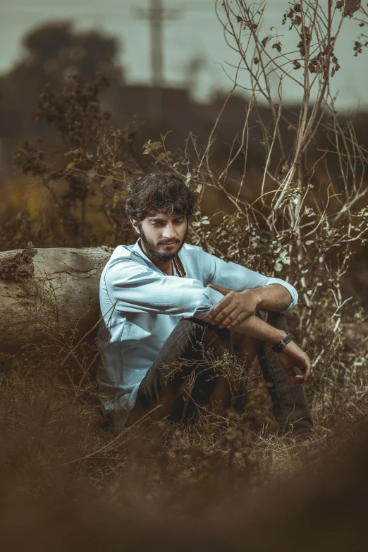 a man sitting in a field next to a tree, a colorized photo, pexels contest winner, renaissance, portrait of a rugged young man, mohamed chahin style, white shirt and blue jeans, scruffy looking