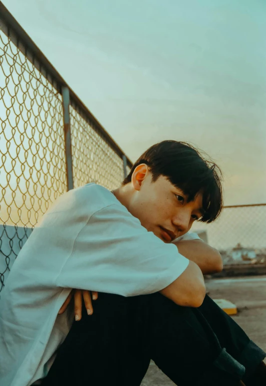 a man sitting on the ground next to a fence, an album cover, inspired by jeonseok lee, unsplash, realism, boyish face, looking off into the sunset, profile image, asian male