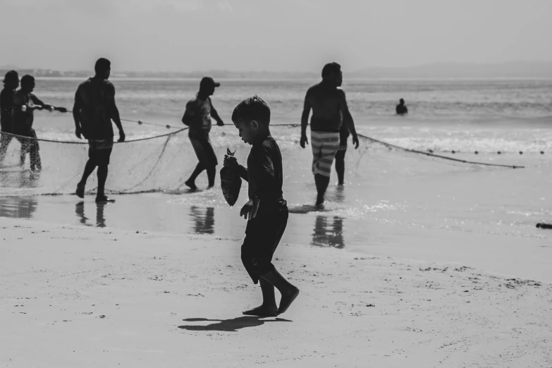 a group of people standing on top of a beach next to the ocean, netting, little boy, monochrome, playing soccer at the beach