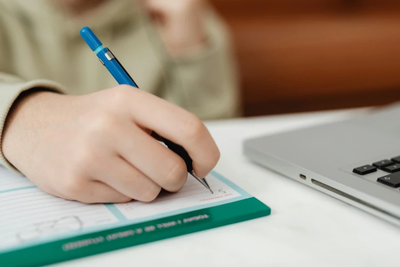 a person writing on a notebook next to a laptop, a child's drawing, pexels, teal paper, blue ballpoint pen, thumbnail, student