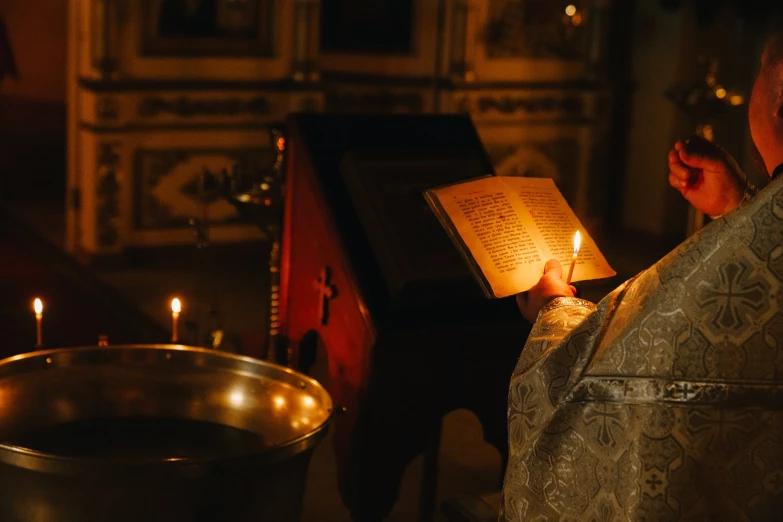 a priest reading a book while holding a lit candle, by Julia Pishtar, pexels, in orthodox church, reading for a party, inside a tomb, basil gogos