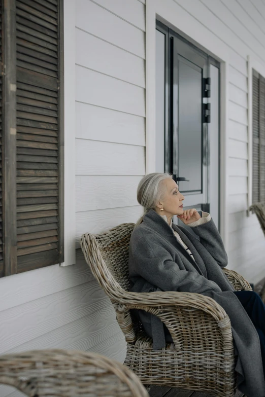 a woman sitting in a wicker chair on a porch, by Matija Jama, pexels contest winner, going gray, shutters, looking away, a cozy