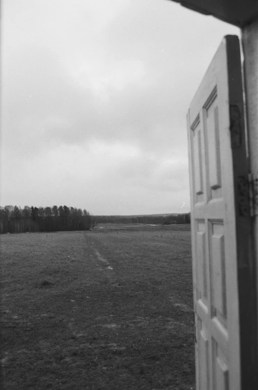 a black and white photo of an open door, inspired by Isaac Levitan, in the middle of a field, distant forest horizon, door to lab, cloudy day