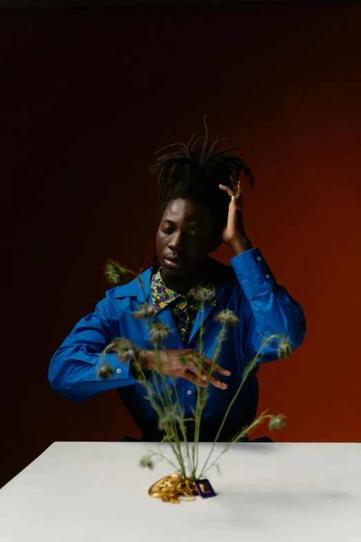 a man sitting at a table with a vase of flowers, an album cover, trending on pexels, dark skinned, expressive pose, indigo, plants