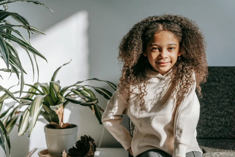 a little girl sitting on a couch next to a potted plant, a character portrait, pexels contest winner, happening, long afro hair, beige hoodie, on a white table, natural lighting