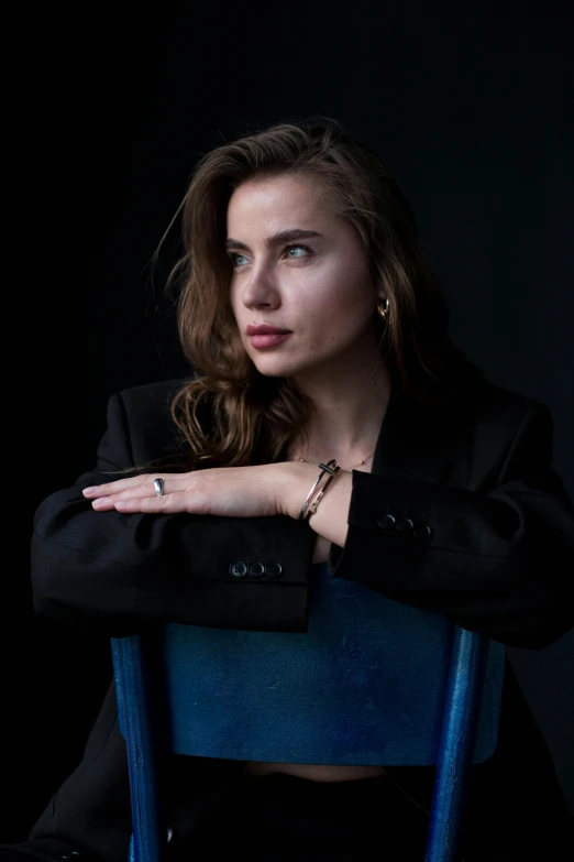 a woman sitting on top of a blue chair, inspired by Elsa Bleda, trending on unsplash, photorealism, wearing jewellery, wearing a black jacket, square, standing with a black background