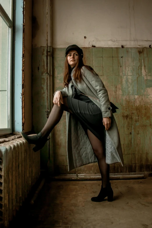 a woman leaning against a radiator in a room, a portrait, inspired by August Sander, pexels contest winner, wearing trenchcoat, tights, in a warehouse, woman with hat