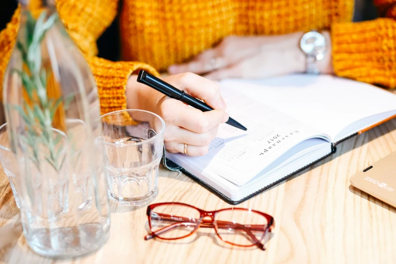 a person sitting at a table writing in a notebook, by Nicolette Macnamara, pexels contest winner, with square glasses, 9 9 designs, drinking, background image