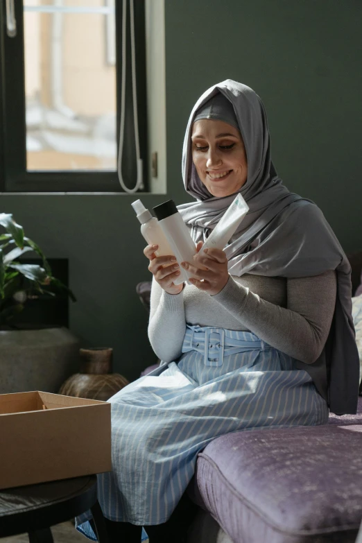 a woman sitting on a couch reading a book, hurufiyya, holding a bottle of arak, inside its box, white hijab, smirking