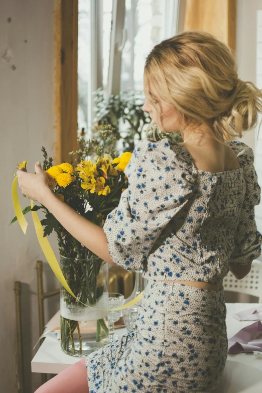 a woman sitting at a table with a vase of flowers, pexels contest winner, lady using yellow dress, yellow and blue ribbons, style of julia razumova, a blond