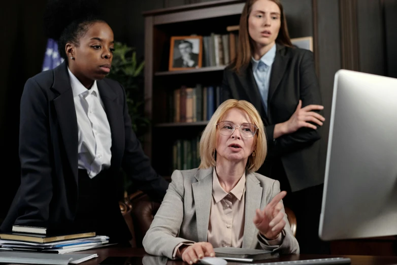 a woman sitting at a desk in front of a computer, pexels, hurufiyya, three women, lawyer suit, group of people in a dark room, a blond