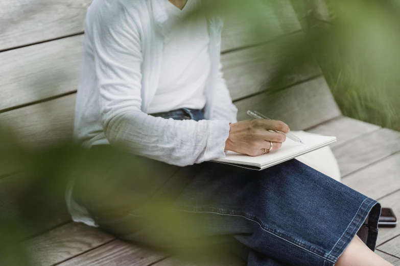 a woman sitting on a bench writing on a notebook, a drawing, by Emma Andijewska, pexels contest winner, white shirt and jeans, amongst foliage, uncropped, clean details