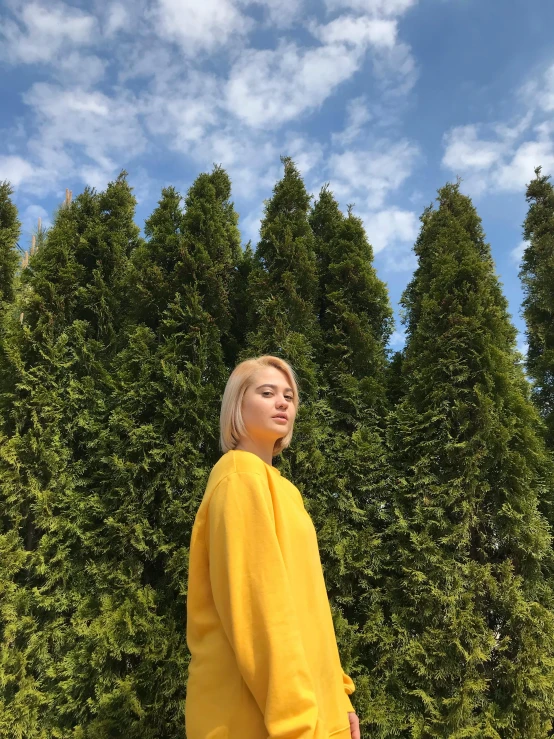 a woman standing in front of a row of trees, by Robbie Trevino, yellow clothes, alexey gurylev, young blonde woman, iphone photo