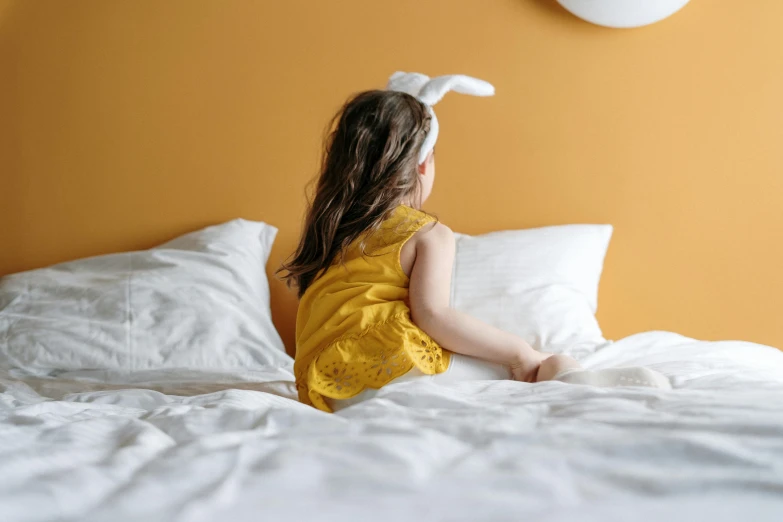 a little girl in a yellow dress sitting on a bed, pexels contest winner, long rabbit ears, facing away, white sheets, 15081959 21121991 01012000 4k