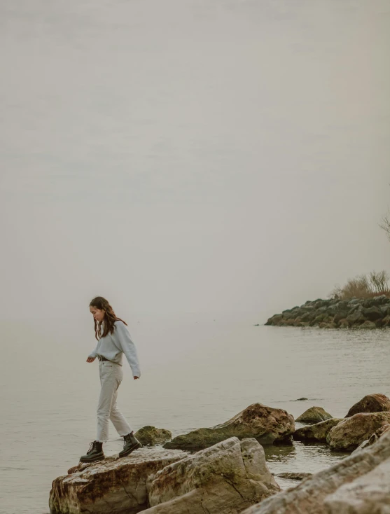 a woman standing on a rock next to a body of water, walking away from the camera, wearing a linen shirt, low quality photo, grey mist