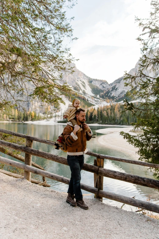 a woman standing on the side of a road next to a lake, father with child, in the dolomites, wooden bridge, a handsome