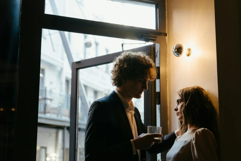 a man and a woman standing in front of a window, by Lee Loughridge, pexels contest winner, flirting, holding a drink, about to enter doorframe, sydney hanson