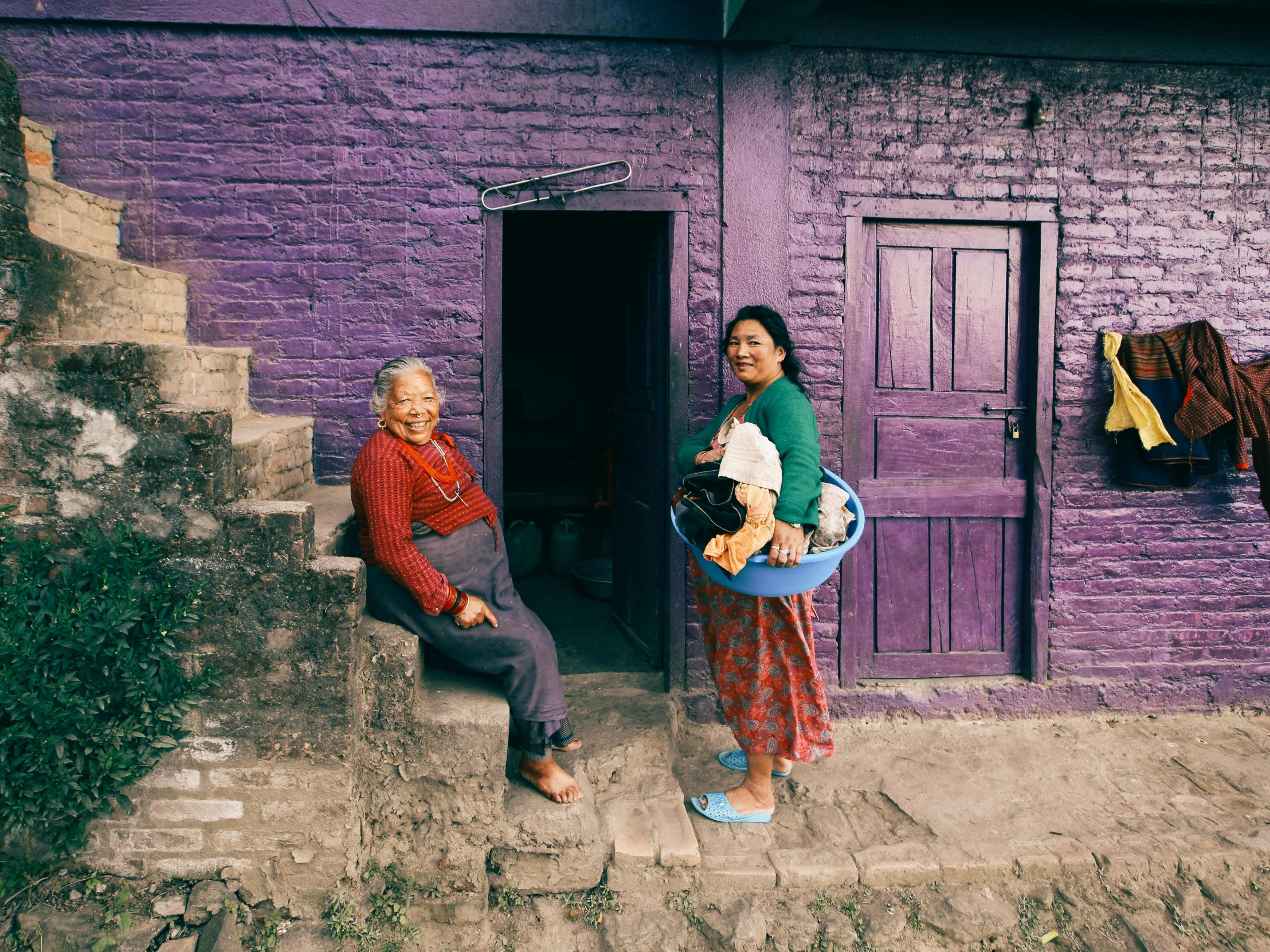 two women standing in front of a purple building, pexels contest winner, cloisonnism, two old people, in the hillside, 2000s photo, nepali architecture buildings