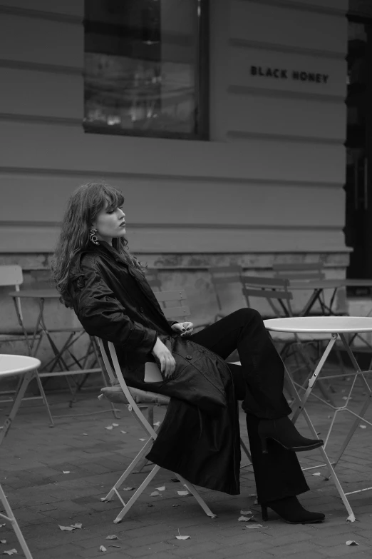 a black and white photo of a woman sitting at a table, inspired by Kati Horna, wearing black leather trenchcoat, on a street, with bangs, siting in a chair