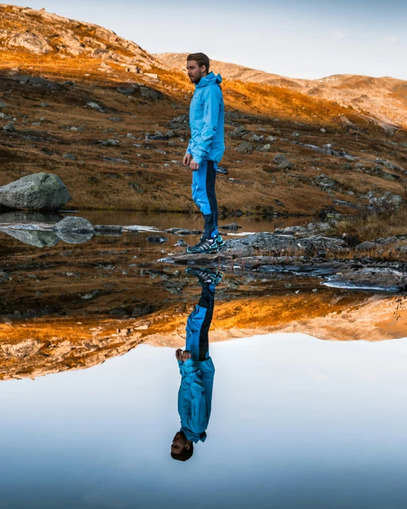 a man standing in front of a body of water, an album cover, by Sebastian Spreng, pexels contest winner, hiking clothes, reflective ground, amber and blue color scheme, norwegian man