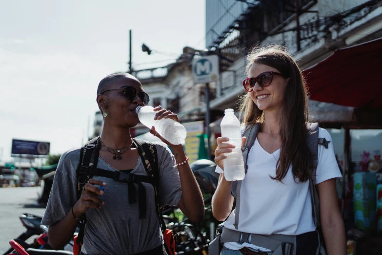 a couple of women standing next to each other on a street, pexels contest winner, hydration, avatar image, water bottles, sweltering heat