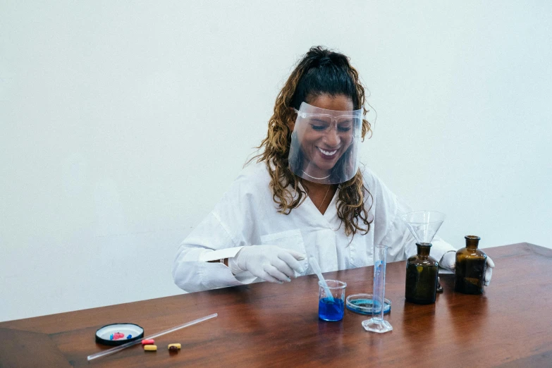 a woman in a lab coat sitting at a table, by Olivia Peguero, pexels contest winner, process art, mutahar laughing, epoxy resin, aida muluneh, wearing transparent glass mask