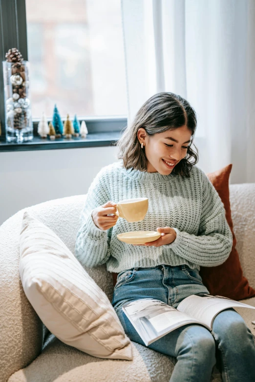 a woman sitting on a couch holding a cup of coffee, trending on pexels, happening, wearing casual sweater, eating, joyful look, reading