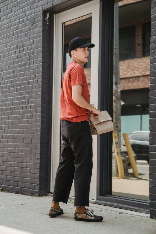 a man standing outside of a building holding a box, matte black pants, holding pizza, red shirt brown pants, storefront
