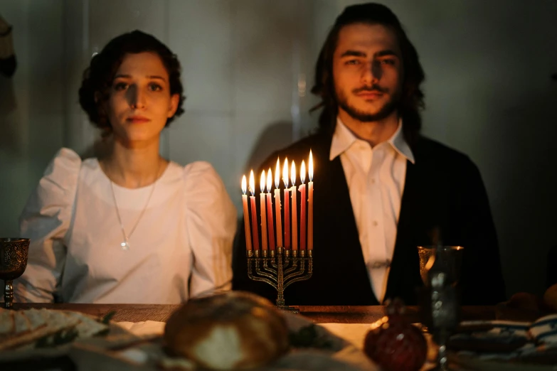 a man and a woman sitting at a table with lit candles, hana alisa omer, square, holiday season, medium-shot