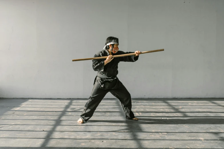 a man holding a wooden stick in a room, inspired by Kanō Hōgai, pexels contest winner, doing a kick, tai costume, black, technical